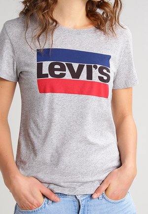 Person iført en grå Levi's t-shirt med en blå og rød rektangulær logo og lyseblå jeans, hænder i lommerne.