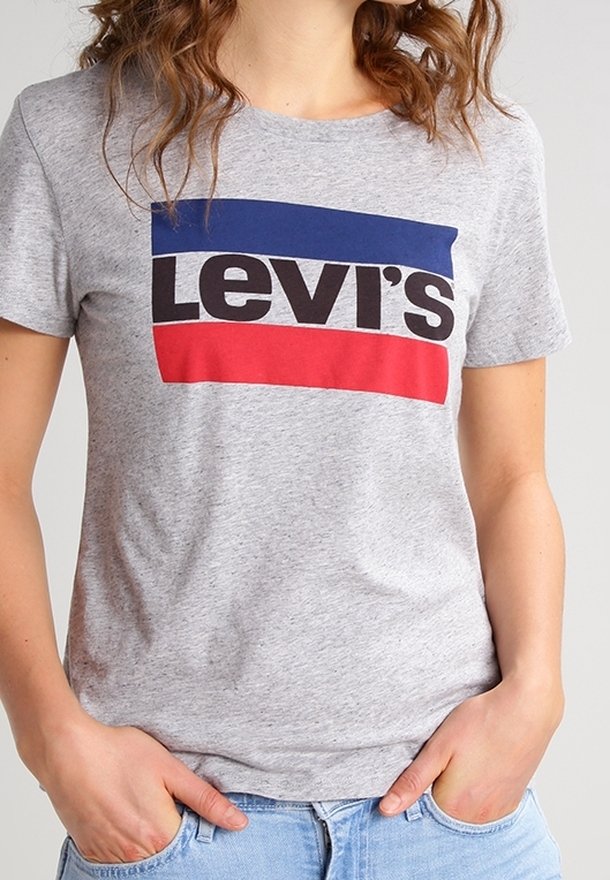 Personne portant un t-shirt gris Levi's avec un logo rectangulaire bleu et rouge et un jean bleu clair, mains dans les poches.