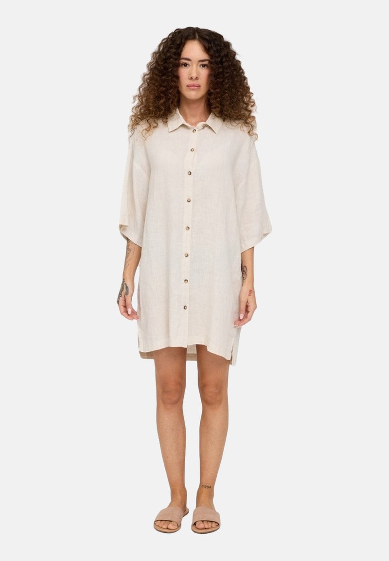 Beiges Leinenhemdkleid mit kurzen Ärmeln, durchgehender Knopfleiste und Seitenschlitzen. Das Model trägt Sandalen und zeigt sichtbare Tattoos an den Armen.