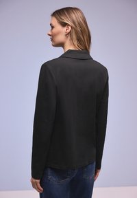 Femme aux cheveux longs blonds portant un haut noir à manches longues et un jean bleu, tournée vers la gauche, de dos à la caméra, sur un fond violet clair.