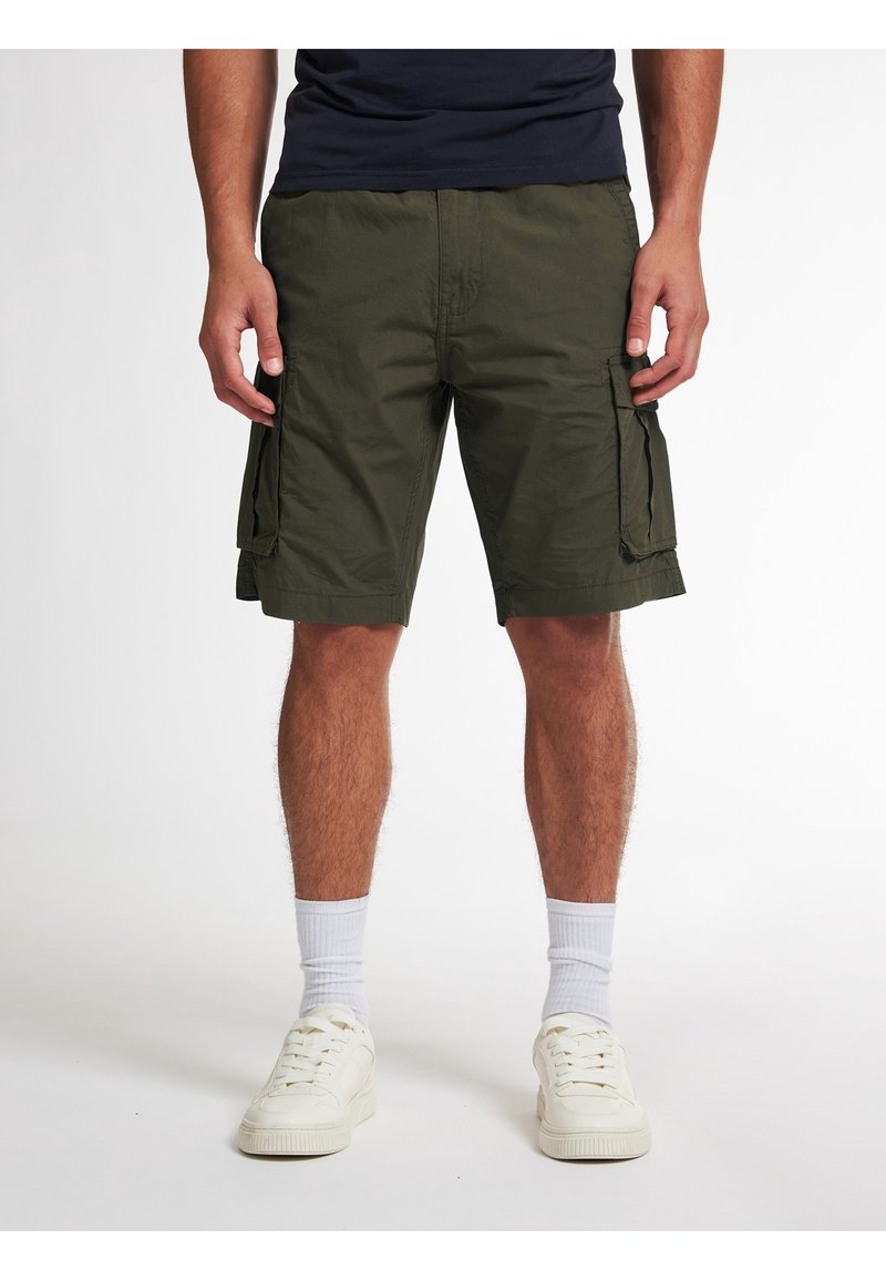 Homme portant un short cargo vert foncé, des chaussettes blanches et des baskets blanches, debout devant un fond blanc uni.