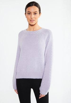 Woman wearing a light purple knitted sweater with small white beads on sleeves and chest, paired with black pants, standing against white background.