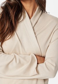 Beżowy sweter o kroju wrap z teksturowanej tkaniny, z dekoltem w serek, długimi rękawami i luźnym krojem. Ręce skrzyżowane z przodu.