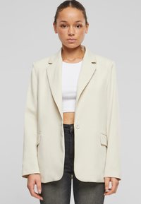 Blazer crema con rever a punta, chiusura a un bottone e due tasche anteriori. Tessuto liscio e vestibilità comoda. Indossato sopra un crop top bianco.