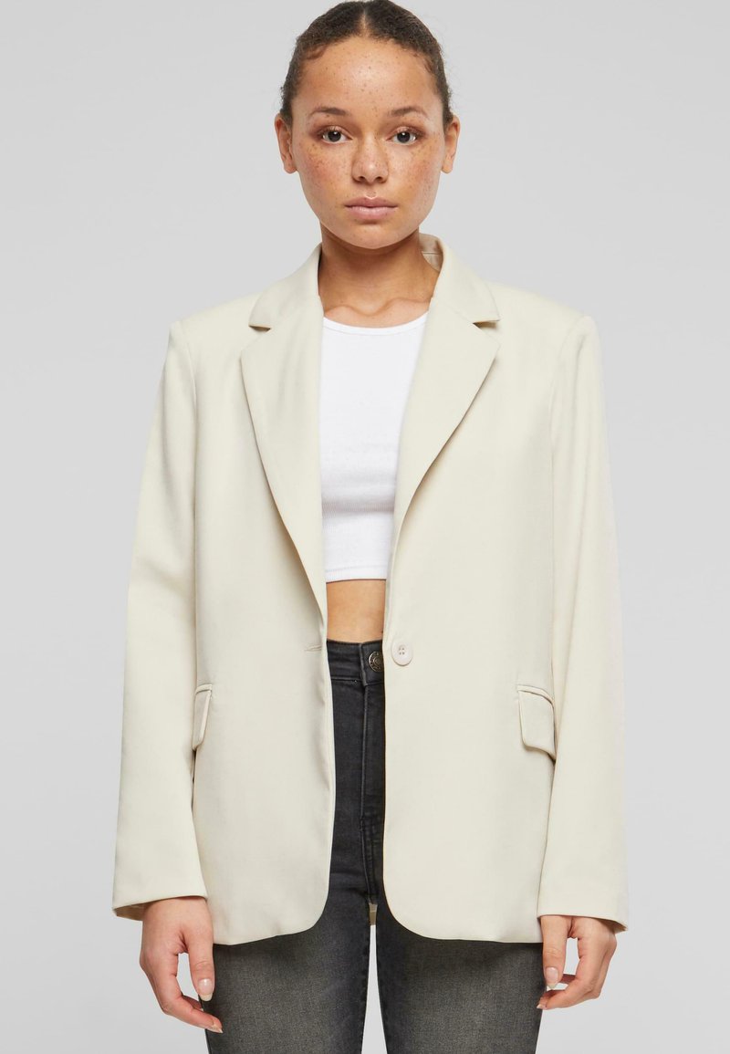 Blazer crema con rever a punta, chiusura a un bottone e due tasche anteriori. Tessuto liscio e vestibilità comoda. Indossato sopra un crop top bianco.