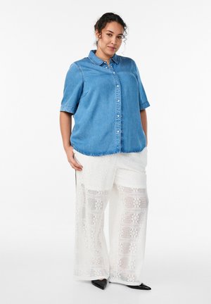 Frau trägt ein blaues Jeanshemd mit kurzen Ärmeln und weiße, weite Spitzenhose, steht mit den Händen in den Taschen vor einem weißen Hintergrund.