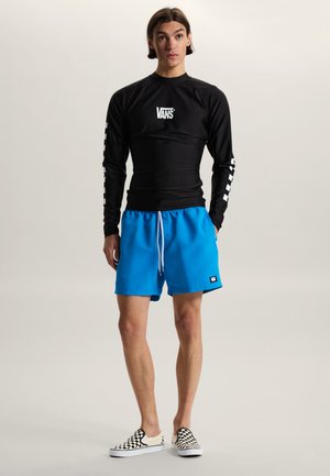 Ung mand står iført sort Vans langærmet skjorte, lyseblå shorts med snøre og sort-hvide ternede slip-on sko.