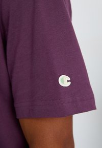 Vijolična bombažna majica s kratkimi rokavi in majhnim belim in zelenim logotipom na levem rokavu. Gladka tekstura in klasična zasnova.