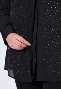 Schwarzes Hemd mit silbernen Verzierungen, mit einer durchgehenden Knopfleiste, langen Ärmeln und einem strukturierten Stoff mit einem leicht transparenten Saum.