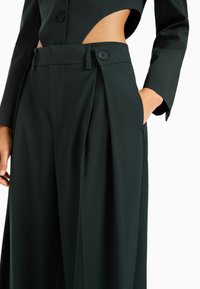 Mörkgröna skräddarsydda byxor med hög midja, veck och sidofickor. Har två svarta knappar vid midjebandet och en mjuk textur.