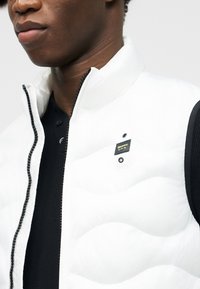 Κοντινό πλάνο ενός ατόμου που φοράει άσπρο γουντριστό γιλέκο με μαύρο φερμουάρ πάνω από μαύρο πουκάμισο, δείχνοντας ένα μικρό μαύρο και κίτρινο λογότυπο.