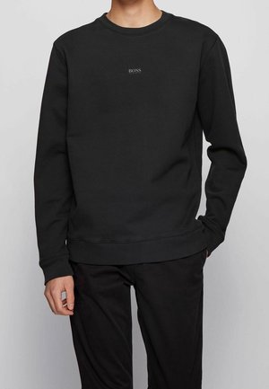 Homme portant un sweat-shirt noir uni à manches longues avec un petit logo « BOSS HUGO BOSS » centré sur la poitrine, associé à un pantalon noir.