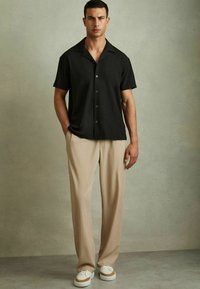 Cămașă neagră cu mânecă scurtă și guler relaxat, asortată cu pantaloni plisati bej și adidași albi cu accente bej.