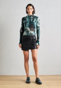 Langermet topp i grønn og teal tie-dye mønster med ribbet tekstur; svart faux lær omslagskjørt med sidestropper; svarte snøresko.