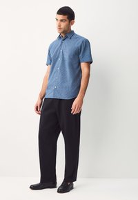 Koszula z krótkim rękawem w niebieski wzór z delikatnym nadrukiem, kołnierzem zapinanym na guziki i kieszenią na piersi, w zestawie z czarnymi spodniami i czarnymi butami.