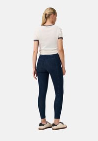 T-shirt bianca con rifiniture grigie, abbinata a leggings in denim scuro. La modella indossa scarpe beige con dettagli neri e suola in gomma.