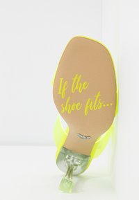 Chaussure à talon haut jaune avec talon transparent, texte sur la semelle indiquant "If the shoe fits…" en écriture cursive jaune sur semelle beige.