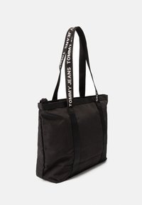 Sac fourre-tout noir en tissu durable, doté de doubles poignées avec le texte "TOMMY JEANS". Le design est minimaliste avec une forme rectangulaire.