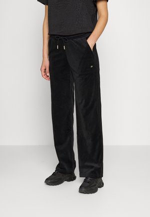 Osoba ubrana w czarną, teksturowaną bluzkę z krótkim rękawem, czarne welurowe spodnie o szerokich nogawkach ze ściągaczem i czarne buty sportowe, stojąca przy białej ścianie.