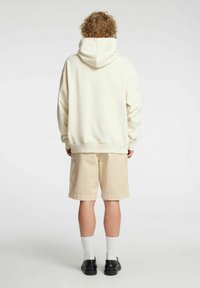 Zgoden kremasto obarvan oversized pulover s kapuco na vrvico, kombiniran z bež kratkimi hlačami. Obleka ima sproščen kroj in subtilne detajle šivov.