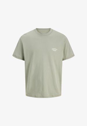 Zelené bavlnené tričko s krátkymi rukávmi a logom na hrudi, skombinované s tmavošedými voľnými džínsami a béžovými mokasínami. Čierna čiapka dokončuje outfit.