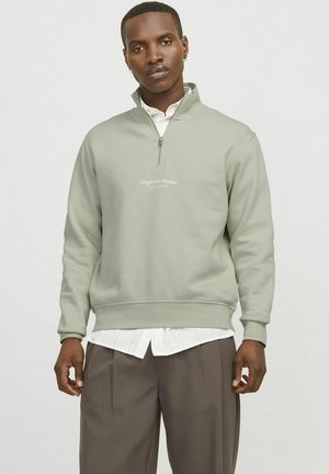 Lichtgroene zip-up sweatshirt van zachte stof, met een hoge kraag, geribbelde manchetten en een geborduurd logo op de borst. Gecombineerd met een wit shirt.