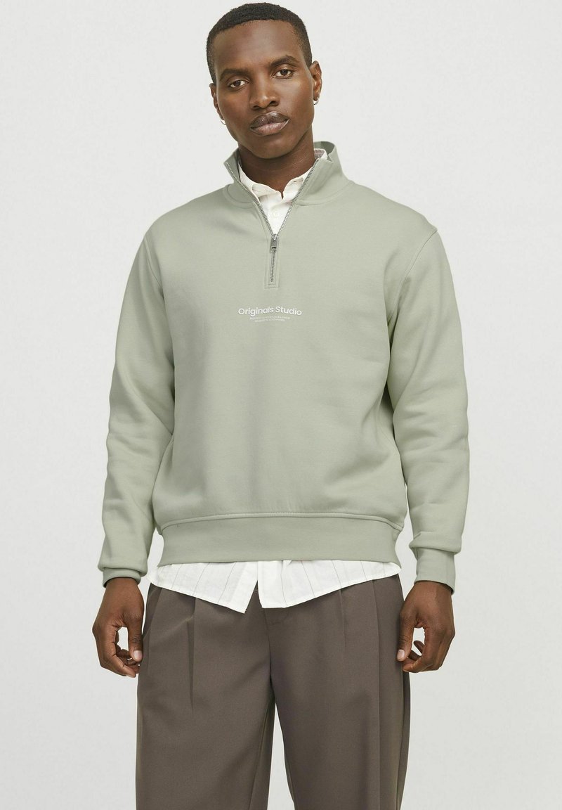 Light green zip-up sweatshirt in soft fabric, featuring a high collar, ribbed cuffs, and an embroidered logo on the chest. Paired with a white shirt.