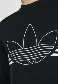 Svart sweatshirt med en stor vit trefoil-logotyp med horisontella ränder och tre vita ränder på ärmarna. Tillverkad av bomull.