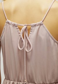 Robe rose clair à fines bretelles, avec un nœud décoratif dans le dos et une taille froncée. Texture lisse avec un léger éclat subtil.