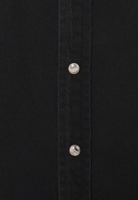 Camisa de tela negra con una línea vertical de botones plateados a lo largo de un lado, que muestra una textura mate y sutiles detalles de costura.