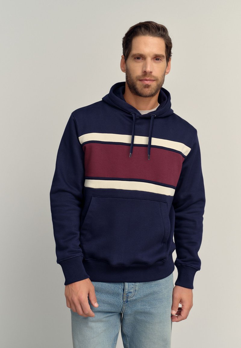 Mann trägt dunkelblauen Hoodie mit weißen und kastanienbraunen horizontalen Streifen, steht vor einfarbig hellem Hintergrund.