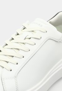 White leather sneaker with beige laces and minimal stitching. Features a rounded toe and a padded collar. Branding visible on the tongue.