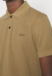 Brun polo-shirt i texturerad tyg, med en placket med två knappar och en liten svart "BOSS"-logotyp på vänster bröst.
