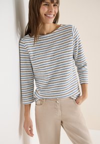 Femme souriante portant un haut en maille rayé bleu, marron et blanc à manches trois-quarts et un pantalon beige, debout devant un mur clair.