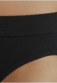 Zwarte ribbelstofdetails van een tailleband, met een gladde textuur en een naadloze rand, waarbij de huidskleur eronder zichtbaar is.