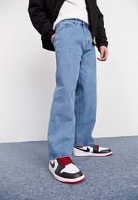 Light blue denim jeans with a straight-leg fit, worn with black, red, and white sneakers featuring a logo, on a textured blue surface.