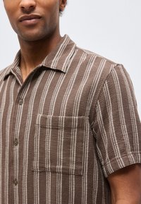 Άνδρας που φοράει καφέ κοντομάνικο πουκάμισο με λευκές κάθετες ρίγες και τσέπη στο στήθος, μπροστά σε απλό φόντο.