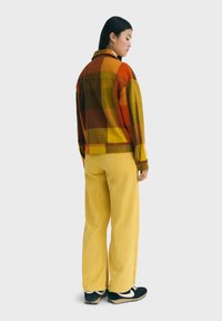 Camisa de franela en cuadros naranjas, amarillos y marrones con un ajuste ligeramente holgado, combinada con pantalones de pana amarillos y zapatillas negras.