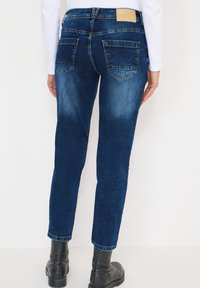 Jean en denim bleu foncé à coupe effilée, avec poches arrière et étiquette en cuir à la ceinture, porté avec des bottines noires.