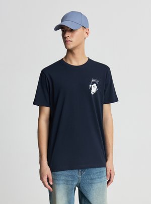 Giovane uomo che indossa una maglietta blu navy con un piccolo personaggio dei cartoni animati che tiene una bandiera sul petto, jeans azzurri e un cappellino da baseball blu.