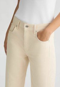 Jeans de talle alto beige con una tela texturizada, bolsillos frontales y cierre de botón plateado, combinados con una camiseta ajustada blanca de canalé.