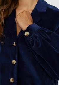 Mornarsko modra kordna srajca s ovratnikom, velikimi gumbi in voluminoznimi rokavi z elastičnimi manšeti. Gladka tekstura in bogata barva.