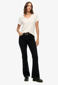 Camisa blanca de cuello en V con mangas cortas, combinada con pantalones vaqueros negros de campana y un cinturón negro con hebilla dorada. El modelo se presenta de forma segura.