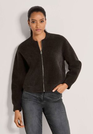 Femme aux cheveux foncés portant une veste noire en peluche à fermeture éclair et un jean gris, debout contre un mur clair avec une main dans la poche.
