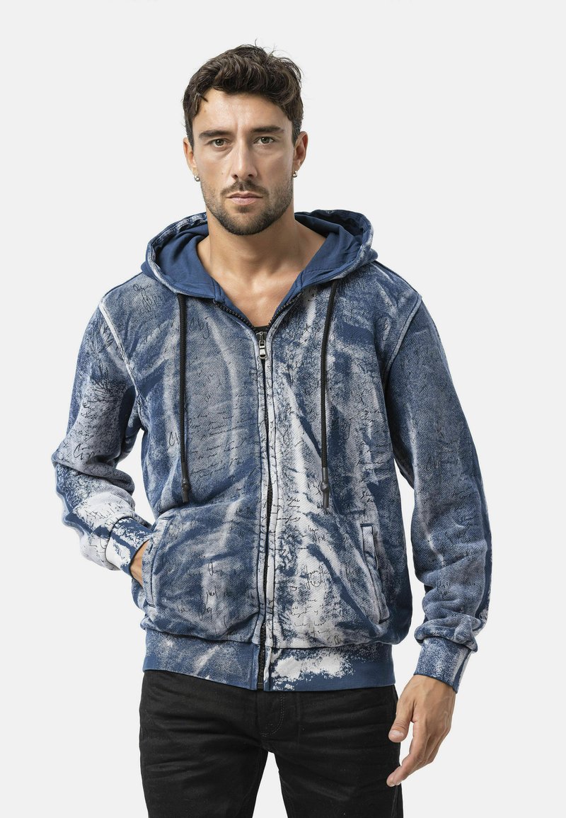 Blauwe hoodie met rits, voorzien van een wit abstract patroon, verstelbare trekkoorden en zijzakken. Gemaakt van een gestructureerde stof voor warmte.