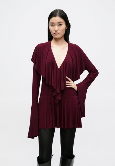 Μπορντό φόρεμα με στρωματοποιημένο σχέδιο με βολάν, V-λαιμόκοψη, μακριά μανίκια καμπάνα και πλισέ φούστα. Το μοντέλο φοράει εφαρμοστά μαύρα κολάν και μπότες μέχρι το γόνατο.