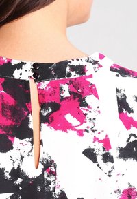 Mehrfarbige Bluse mit einem abstrakten Muster in Schwarz, Weiß und Pink. Sie hat eine Schlüsselchenöffnung am Rücken mit zwei schwarzen Knöpfen.