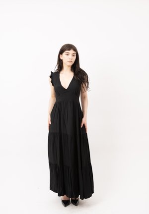 Femme aux longs cheveux foncés portant une robe longue noire sans manches à volants et des chaussures noires pointues, debout devant un fond blanc.