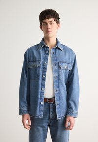 Lys blå denimskjorte med to brystlommer, knappelukking og klassisk krage. Båret over en hvit T-skjorte med et brunt belte.