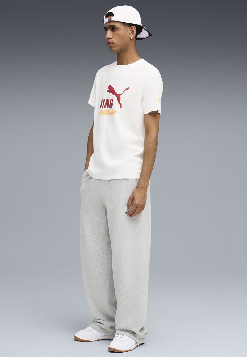 Camiseta blanca con logo de Puma en rojo y amarillo, combinada con pantalones de chándal grises holgados y zapatillas blancas. Gorra ajustable blanca llevada al revés.